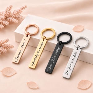 May include: Four rectangular metal keychains with circular rings. The first is rose gold with 'Jeano, Jose!' engraved. The second is gold with 'infinitely & always'. The third is black with 'Your Handwriting'. The fourth is silver with 'Un jour à la fois'.