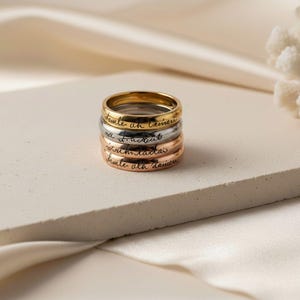 Peut inclure: Une pile de quatre bagues en m&eacute;tal aux tons or, argent et or rose. Chaque bague pr&eacute;sente un anneau lisse et arrondi avec une inscription en &eacute;criture cursive noire grav&eacute;e sur la surface ext&eacute;rieure. Les bagues sont empil&eacute;es verticalement.