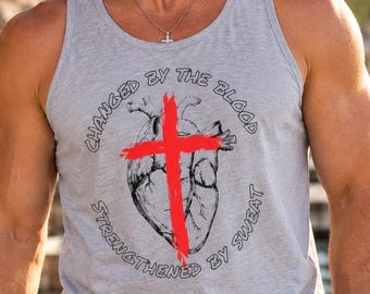 May include: A light gray tank top featuring a black anatomical heart illustration overlaid with a bold red cross. The text "CHANGED BY THE BLOOD" is arched above the heart, and "STRENGTHENED BY SWEAT" is arched below in black lettering.