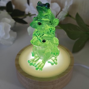 May include: A stack of three translucent lime green frogs made of resin, positioned on a circular light-up wooden base. The frogs are stacked vertically, with each one featuring detailed textures and dark eyes, illuminated from below by the base.