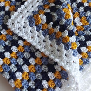 Peut inclure: Une couverture pour bébé en crochet dans des tons de bleu, gris et jaune avec une bordure en crochet blanc.