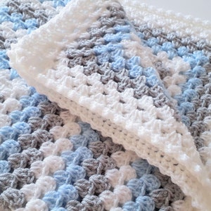 May include: A crocheted baby blanket in shades of blue, gray, and white. The blanket is made with a textured stitch and has a soft, fluffy feel.