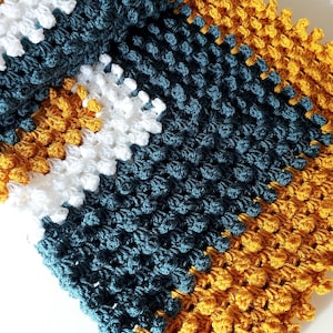 Könnte beinhalten: Eine gehäkelte Decke mit einem gestreiften Muster in Teal, Weiß und Senfgelb. Die Decke ist mit einem strukturierten Stich gearbeitet, der eine unebene Oberfläche erzeugt.