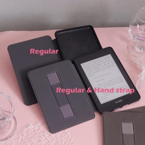 K&ouml;nnte beinhalten: Eine Auswahl an grauen Kindle E-Reader-H&uuml;llen. Eine H&uuml;lle ist ge&ouml;ffnet und zeigt den Kindle-Bildschirm mit Text. Die H&uuml;llen umfassen einen regul&auml;ren Stil und einen regul&auml;ren Stil mit Handschlaufe. Das Wort "Kindle" ist auf dem Ger&auml;t sichtbar.
