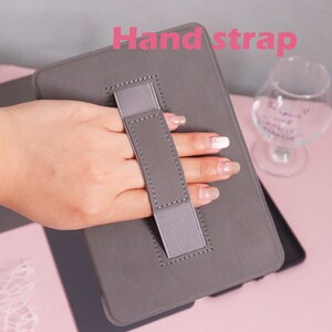 K&ouml;nnte beinhalten: Graue Tablet-H&uuml;lle mit Handschlaufe. Die H&uuml;lle hat ein gen&auml;htes Design und einen grauen Stoffriemen zum Halten. Der Text "Hand strap" ist in Pink sichtbar.