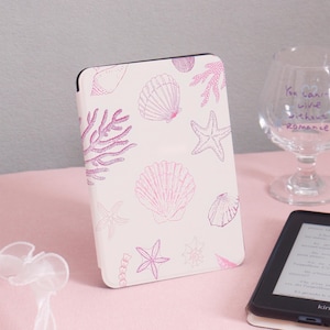 Könnte beinhalten: Eine weiße E-Reader-Hülle mit einem rosa Muschel- und Korallenmuster. Die Hülle zeigt verschiedene Muschelillustrationen, darunter Seesterne und Korallen, in Rosatönen. Die Hülle wird neben einem Kindle E-Reader gezeigt.