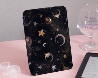 Könnte beinhalten: Schwarze E-Reader-Hülle mit einem Himmelsdesign mit goldenen Sternen, Monden und Punkten. Die Hülle hat einen dunklen Hintergrund mit Aquarelleffekt. Die Hülle liegt auf einer rosa Oberfläche.