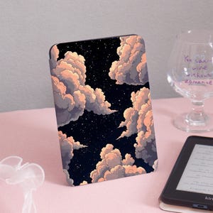 May include: A rectangular case featuring a dreamy night sky design with fluffy, peach-colored clouds against a dark blue background. The case is likely for an e-reader or tablet, with a black border. A glass with text is in the background.