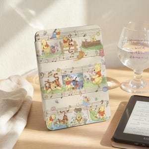 May include: A beige e-reader cover featuring a whimsical design of Winnie the Pooh and friends, including Tigger, Piglet, and Eeyore, set against a musical staff background. The cover is on a wooden surface, next to a glass of water and an e-reader.