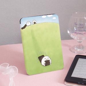 Puede incluir: Una funda verde y azul con una ilustración de dibujos animados de bolas de arroz onigiri. La funda tiene un fondo verde claro con un diseño de colinas y un cielo azul. Los personajes onigiri sonríen.