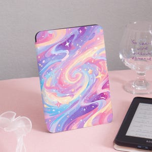 Puede incluir: Una funda rectangular para lector electrónico con un diseño colorido y arremolinado en tonos rosa, morado, azul y amarillo. La funda presenta un patrón de galaxia con estrellas. La funda está sobre una superficie rosa.