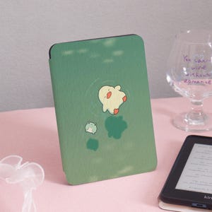 May include: Green e-reader case with a whimsical design featuring a cartoon creature and a frog. The case has a matte finish and is designed to protect an e-reader. The case is shown on a pink surface.