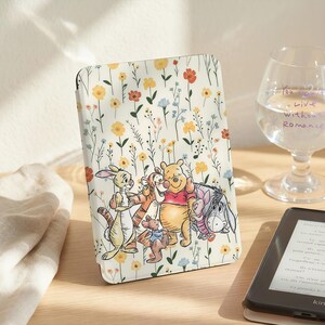 May include: A cream-colored e-reader case featuring a floral pattern with yellow, orange, and blue flowers. The case has a colorful illustration of Winnie the Pooh and friends. The case is on a wooden surface.