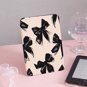 May include: A light pink e-reader case featuring a pattern of black bows. The bows are stylized with a brushstroke effect. The case is standing upright on a pink surface.