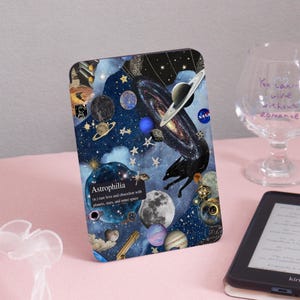 May include: A decorative case with a space-themed design, including planets, stars, and galaxies. The case features the text "Astrophilia" and "NASA" logos, along with a black wolf silhouette. It is shown on a pink surface.