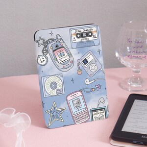 Puede incluir: Una funda azul claro con un diseño retro que presenta una cinta de cassette, teléfonos móviles, un CD y un reproductor de MP3. La funda tiene el texto "SLOW DANCING" y "ANGTAN MUSIC". Un lector Kindle y un vaso con texto están en el fondo.