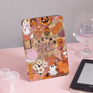 Peut inclure: Une coque décorative pour liseuse avec un collage sur le thème d'Halloween. Le design comprend des citrouilles, des squelettes, des fantômes et les mots "Autumn" et "Spooky Vibes". L'étui est posé sur une surface rose.