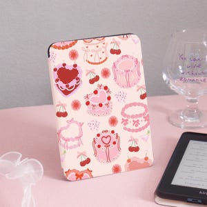 May include: A decorative case for an e-reader featuring a repeating pattern of pink cakes, cherries, and flowers on a light pink background. The case is rectangular and has a black border. A glass with text is in the background.