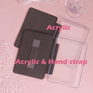 K&ouml;nnte beinhalten: Eine braune Tablet-H&uuml;lle aus Acryl mit Handschlaufe und einer transparenten Acrylabdeckung. Die Texte "Acrylic" und "Acrylic & Hand strap" sind sichtbar. Die H&uuml;lle liegt auf einer rosa Oberfl&auml;che.