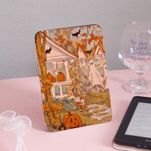 Może przedstawiać: Etui na czytnik e-booków z motywem Halloween. Zawiera ilustrację nawiedzonego domu, dyni, duchów i nietoperzy w jesiennych kolorach. Design ma styl vintage, tworząc upiorną i świąteczną atmosferę.