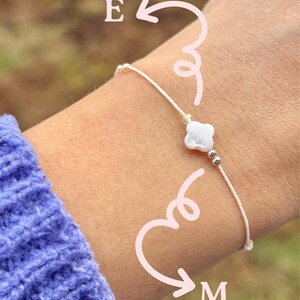 May include: A white string bracelet with a white clover charm and silver beads. The bracelet is on a person's wrist.