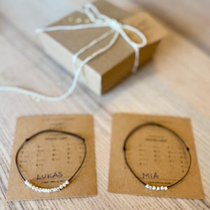 May include: Two personalized bracelets with black string and silver beads spelling out the names "Lukas" and "Mia". The bracelets are displayed on brown cards with white text.