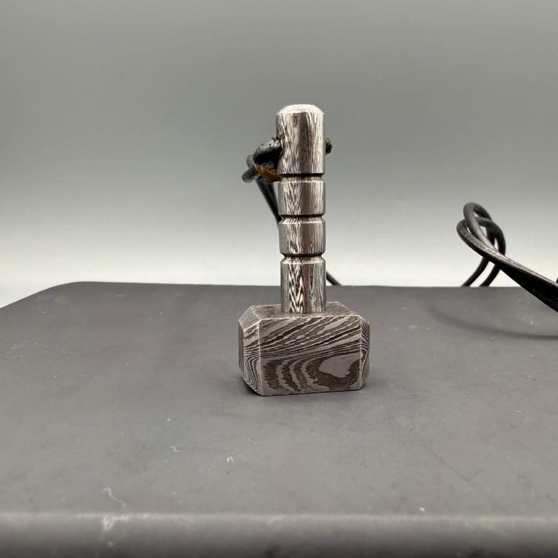Puede incluir: Un peque&ntilde;o colgante de martillo plateado con un cord&oacute;n de cuero negro. La cabeza del martillo tiene forma cuadrada con un patr&oacute;n de vetas de madera. El mango es cil&iacute;ndrico y segmentado. El colgante es probablemente una joya.