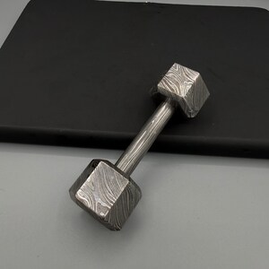 May include: A small, decorative dumbbell with a patterned, textured surface. The dumbbell has two square-shaped weights connected by a cylindrical handle. The metal has a unique, swirling design.