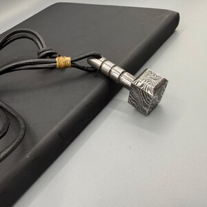 Puede incluir: Un collar con colgante plateado con un dise&ntilde;o en forma de martillo. La cabeza del martillo tiene una textura estampada &uacute;nica, y el collar tiene un cord&oacute;n de cuero negro. El cord&oacute;n est&aacute; asegurado con hilo dorado.