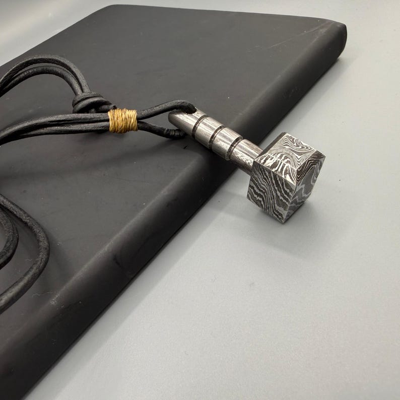 Puede incluir: Un collar con colgante plateado con un dise&ntilde;o en forma de martillo. La cabeza del martillo tiene una textura estampada &uacute;nica, y el collar tiene un cord&oacute;n de cuero negro. El cord&oacute;n est&aacute; asegurado con hilo dorado.