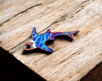 Puede incluir: Un llavero con forma de tiburón con un vibrante patrón azul y morado, acentuado con tonos dorados y plateados. El llavero de metal está sobre una superficie de madera envejecida, mostrando su diseño único y artesanía.