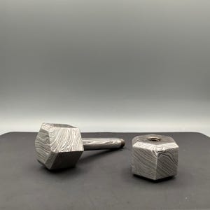 May include: A hammer-shaped bottle stopper and a matching hexagonal bottle stopper. Both are made of a patterned, textured metal with a silver finish. The hammer has a long handle and a large, multi-faceted head.
