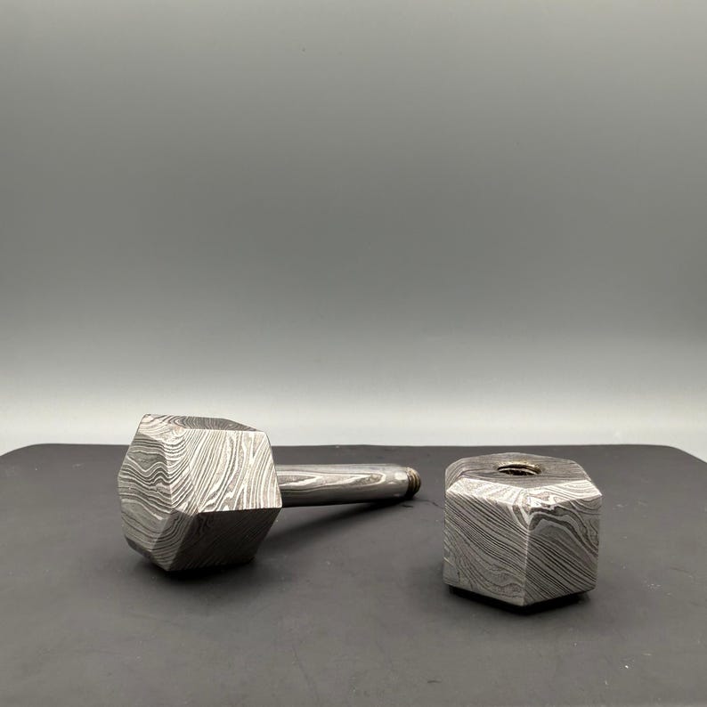 May include: A hammer-shaped bottle stopper and a matching hexagonal bottle stopper. Both are made of a patterned, textured metal with a silver finish. The hammer has a long handle and a large, multi-faceted head.