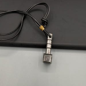 Puede incluir: Un collar con colgante plateado con una base cuadrada y una parte superior cil&iacute;ndrica. El colgante est&aacute; suspendido de un cord&oacute;n de cuero negro. La base cuadrada del colgante presenta un patr&oacute;n distintivo.