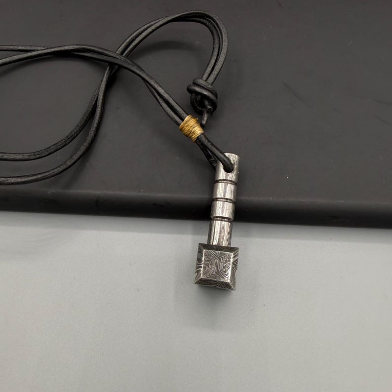 Puede incluir: Un collar con colgante plateado con una base cuadrada y una parte superior cil&iacute;ndrica. El colgante est&aacute; suspendido de un cord&oacute;n de cuero negro. La base cuadrada del colgante presenta un patr&oacute;n distintivo.