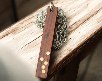 Puede incluir: Un colgante rectangular de madera con cuatro puntos de latón cuelga de una cadena plateada. El colgante es de un color marrón oscuro intenso, y los puntos de latón proporcionan un acento metálico contrastante. La cadena es de diseño clásico.