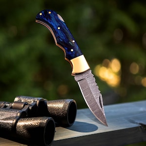 May include: A folding knife with a patterned blade and a blue and gold handle. The handle has a dark blue and green swirl design with gold accents. The blade has a unique pattern. The knife is shown with a pair of binoculars.