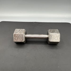 May include: A dumbbell with a patterned, textured surface. The dumbbell has a silver handle and two octagonal weights on each end. The weights have a unique, swirling pattern. The dumbbell is designed for weightlifting and strength training.