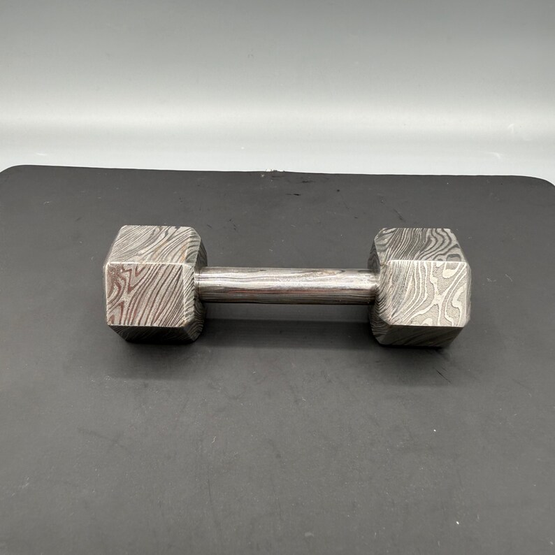 May include: A dumbbell with a patterned, textured surface. The dumbbell has a silver handle and two octagonal weights on each end. The weights have a unique, swirling pattern. The dumbbell is designed for weightlifting and strength training.