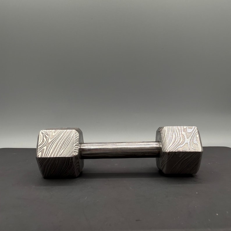 May include: A dumbbell with a patterned design on the ends and a smooth, silver-colored handle. The ends have a unique, textured appearance, and the dumbbell is set against a neutral background. This fitness equipment is designed for strength training.