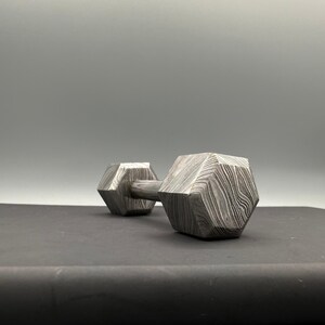 May include: A patterned metal dumbbell with a geometric design. The dumbbell has a hexagonal shape on each end, connected by a cylindrical handle. The metal has a unique, swirling pattern, and the dumbbell is set against a gray background.