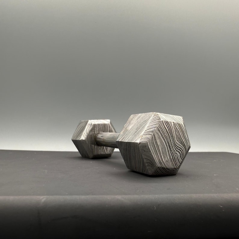 May include: A patterned metal dumbbell with a geometric design. The dumbbell has a hexagonal shape on each end, connected by a cylindrical handle. The metal has a unique, swirling pattern, and the dumbbell is set against a gray background.