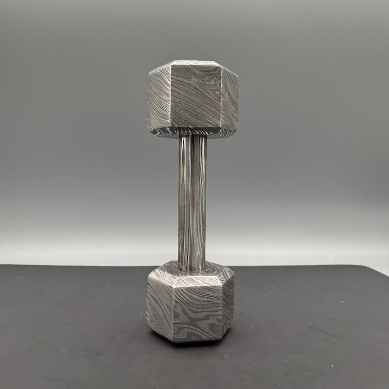 May include: A silver dumbbell with a unique patterned design. The dumbbell has a hexagonal shape on each end, connected by a cylindrical handle. The surface has a textured, swirling pattern.