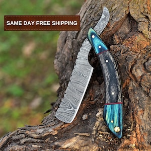 May include: A straight razor with a blue and black handle and a damascus steel blade. The blade has a serrated edge and is partially visible. The handle has a red accent and a gold accent. The razor is resting on a tree trunk.