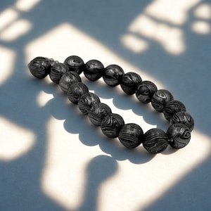 Könnte beinhalten: Ein dunkelgraues Perlenarmband mit einem einzigartigen Muster. Das Armband besteht aus runden Perlen mit einer wirbelnden, holzmaserungsähnlichen Textur. Die Perlen sind zu einem vollständigen Kreis aufgereiht.