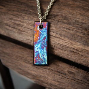 May include: A rectangular pendant necklace with a silver chain. The pendant features a vibrant, abstract design with shades of orange, blue, and purple. The pendant is set against a dark wooden background, highlighting the colorful design.