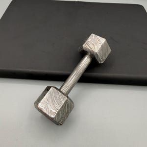 May include: A small, dumbbell-shaped object made of patterned metal. The dumbbell has two square-shaped weights connected by a cylindrical handle. The metal has a unique, swirling pattern, and the object is resting on a dark surface.