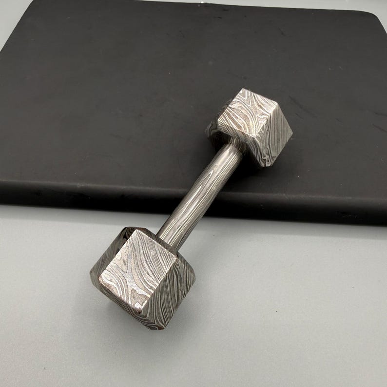 May include: A small, dumbbell-shaped object made of patterned metal. The dumbbell has two square-shaped weights connected by a cylindrical handle. The metal has a unique, swirling pattern, and the object is resting on a dark surface.
