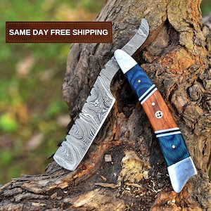 May include: A straight razor with a blue and brown handle and a Damascus steel blade. The razor is lying on a piece of wood.