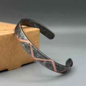 May include: A silver bracelet with a patterned design and copper accents. The bracelet is made of metal and has a smooth finish.